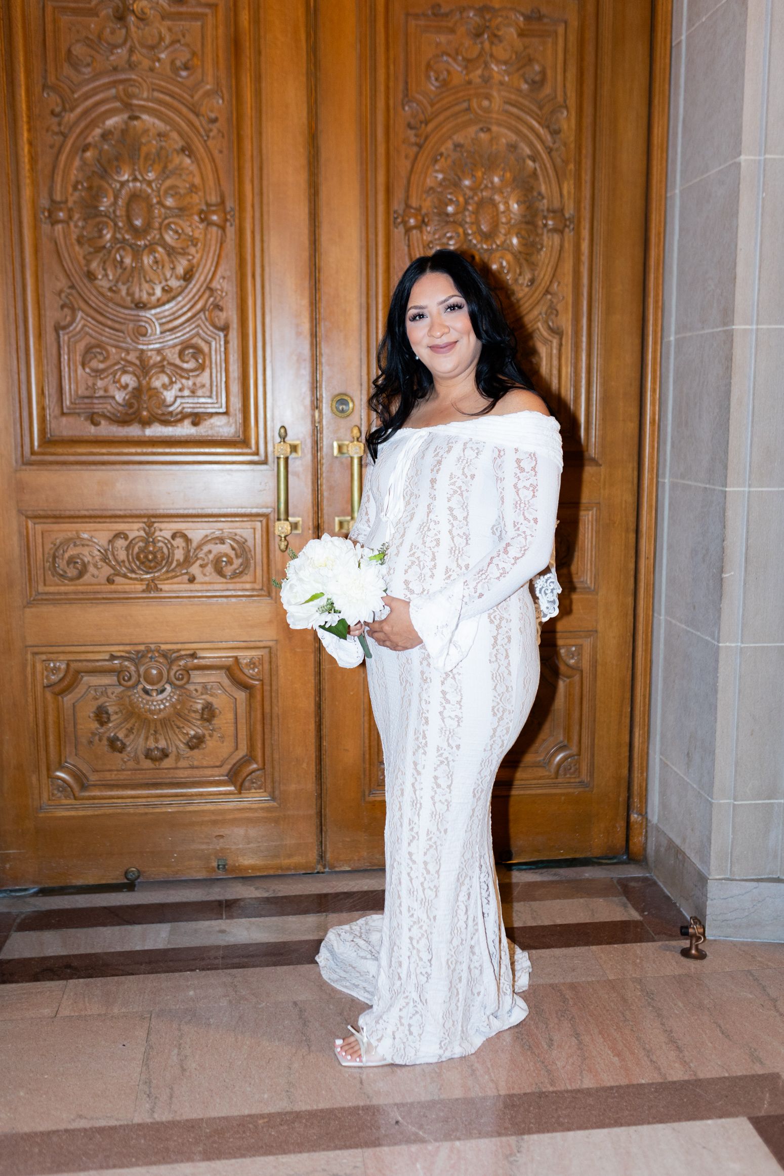 Bride in white off-shoulder lace gown holding bouquet of white roses, posing before ornate wooden doors.