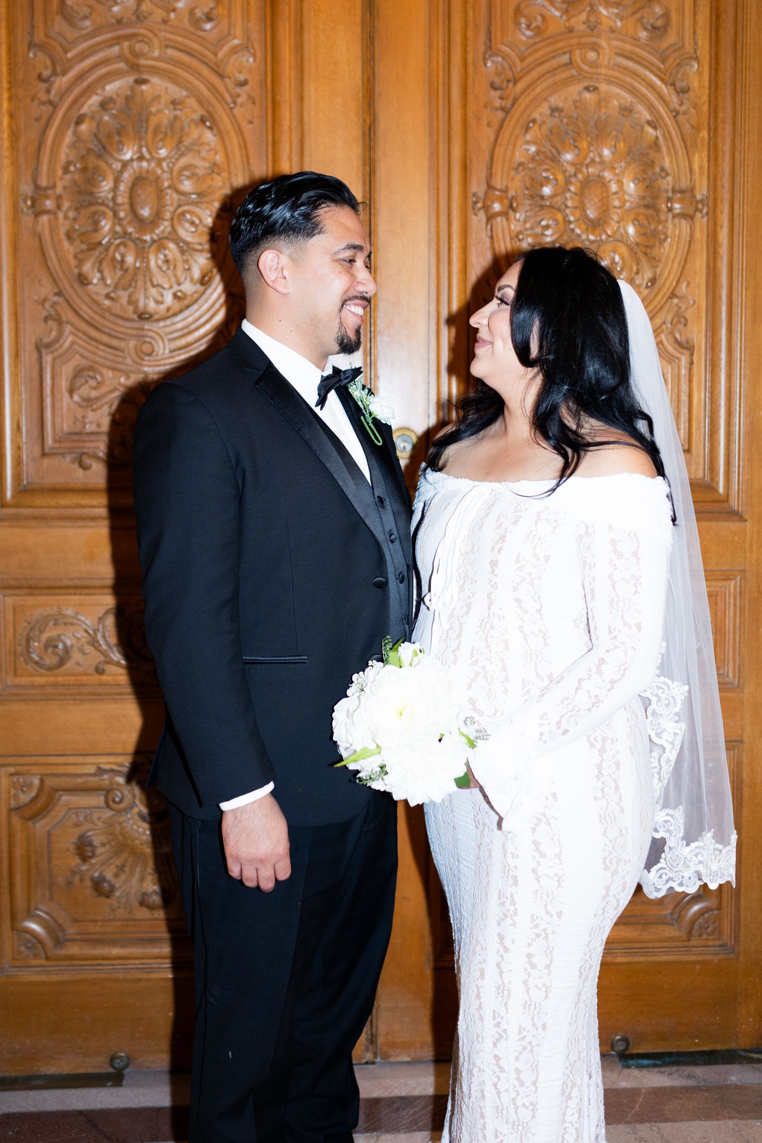 A bride in a white lace gown and groom in a black tuxedo share a joyful moment before ornate wooden doors.