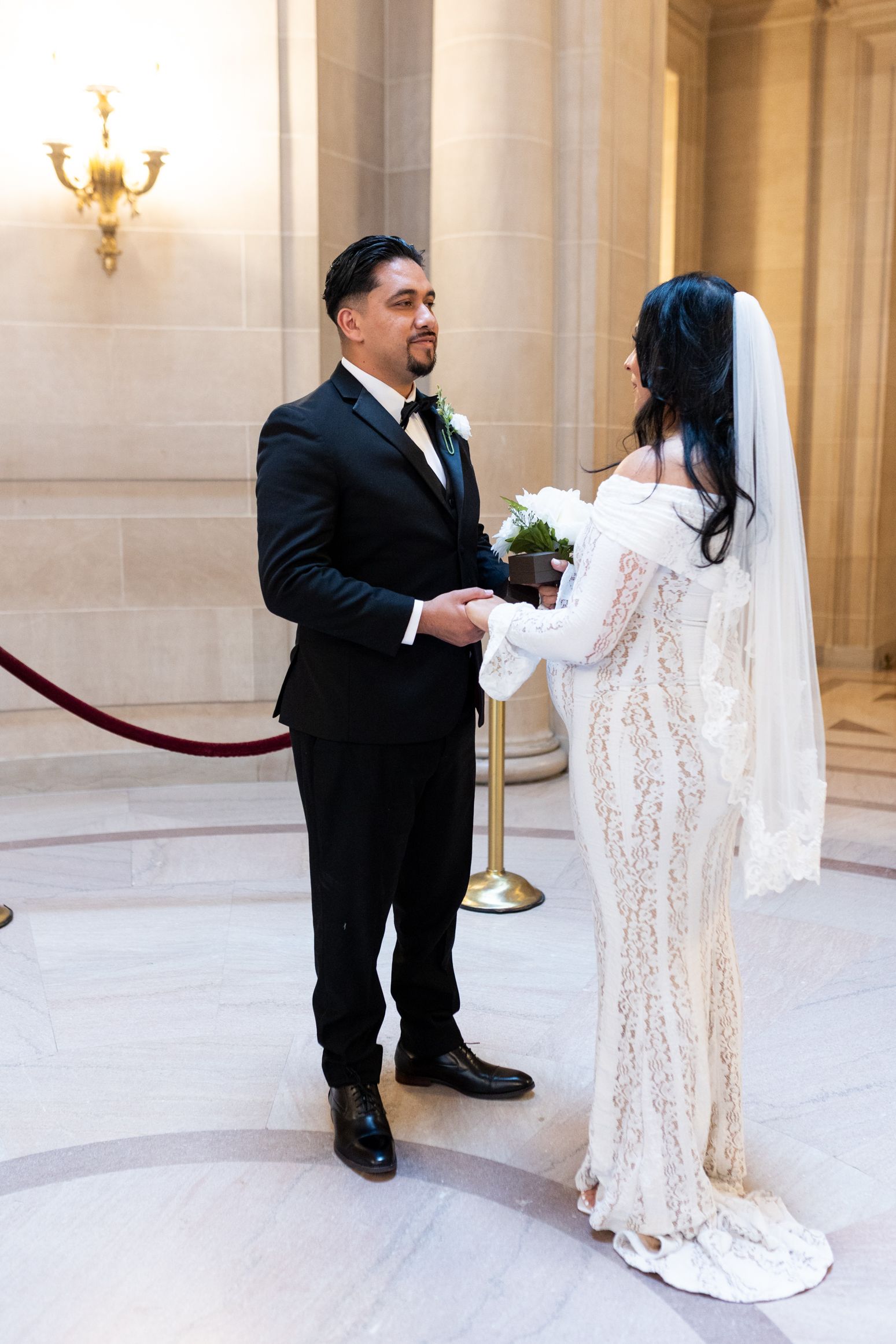 Bride in lace gown and groom in black suit exchange vows at elegant San Francisco City Hall wedding ceremony.