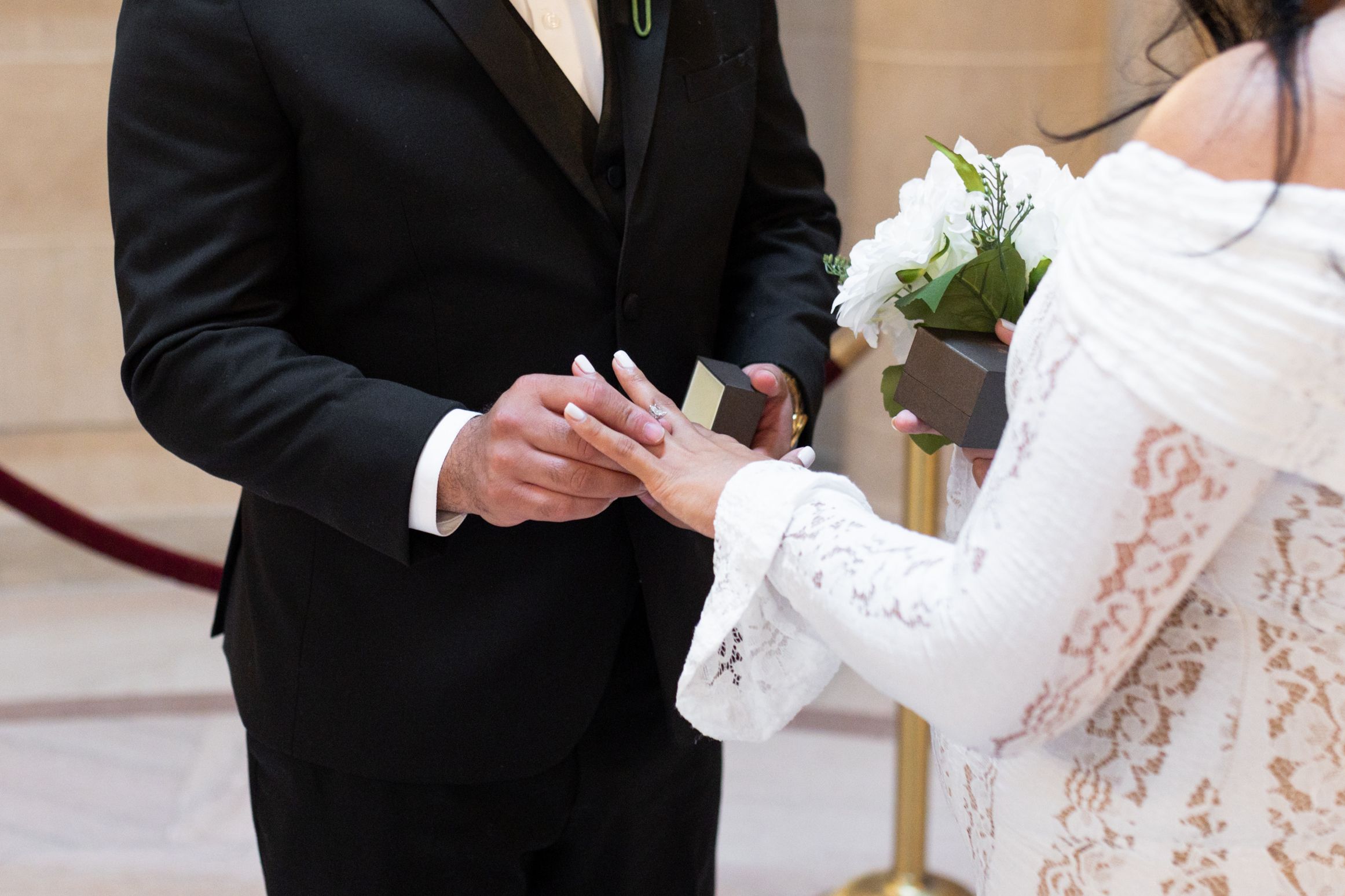 Groom placing a wedding ring on bride's finger during ceremony, bride holding white flowers in elegant lace dress.