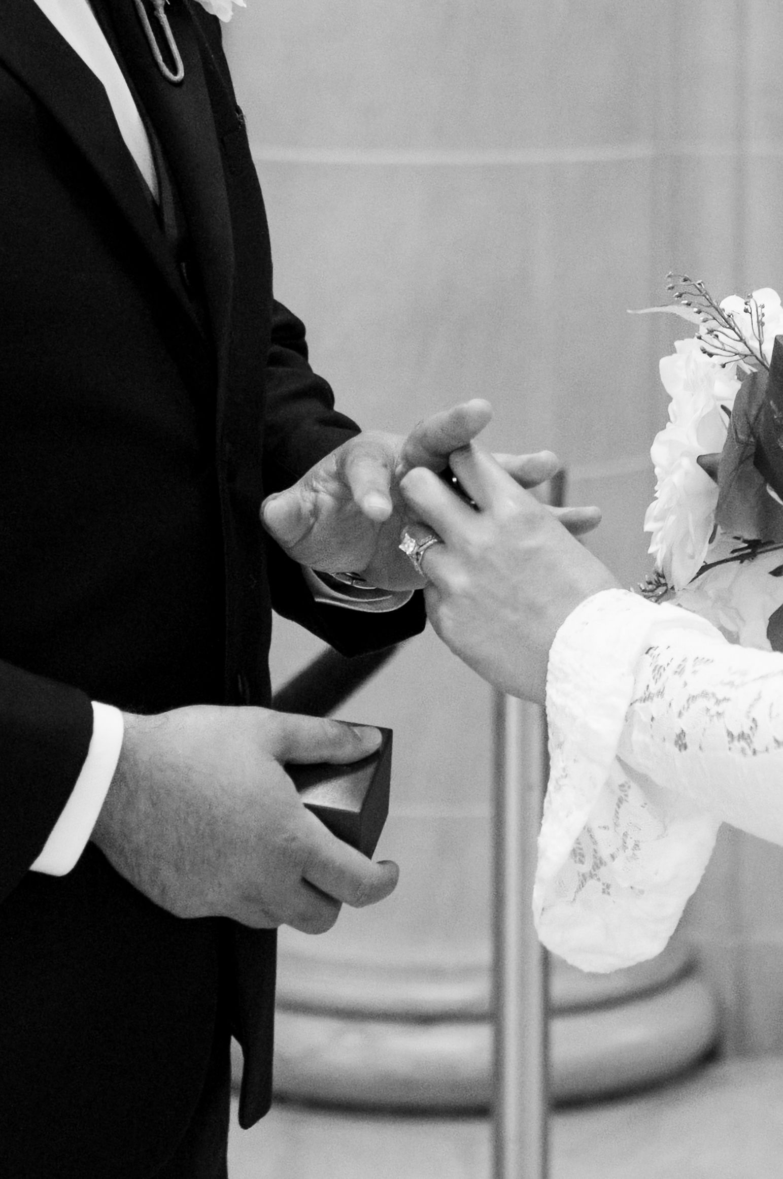 Black and white photo of a couple exchanging wedding rings during a ceremony, bride holding flowers.