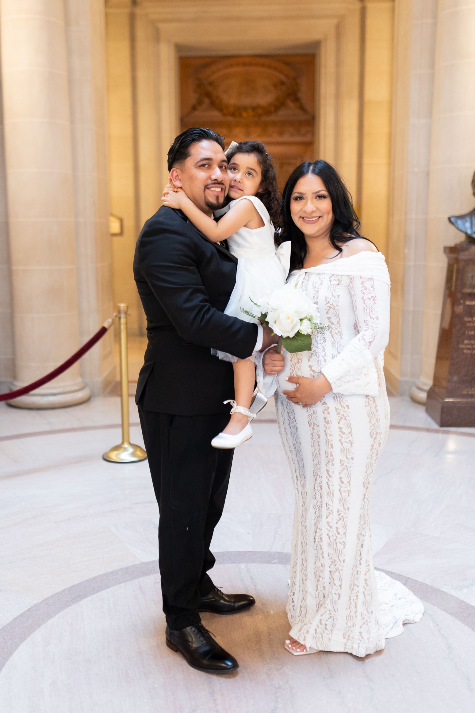 A couple in wedding attire holds their young daughter at an elegant indoor venue, the bride carrying a white bouquet.