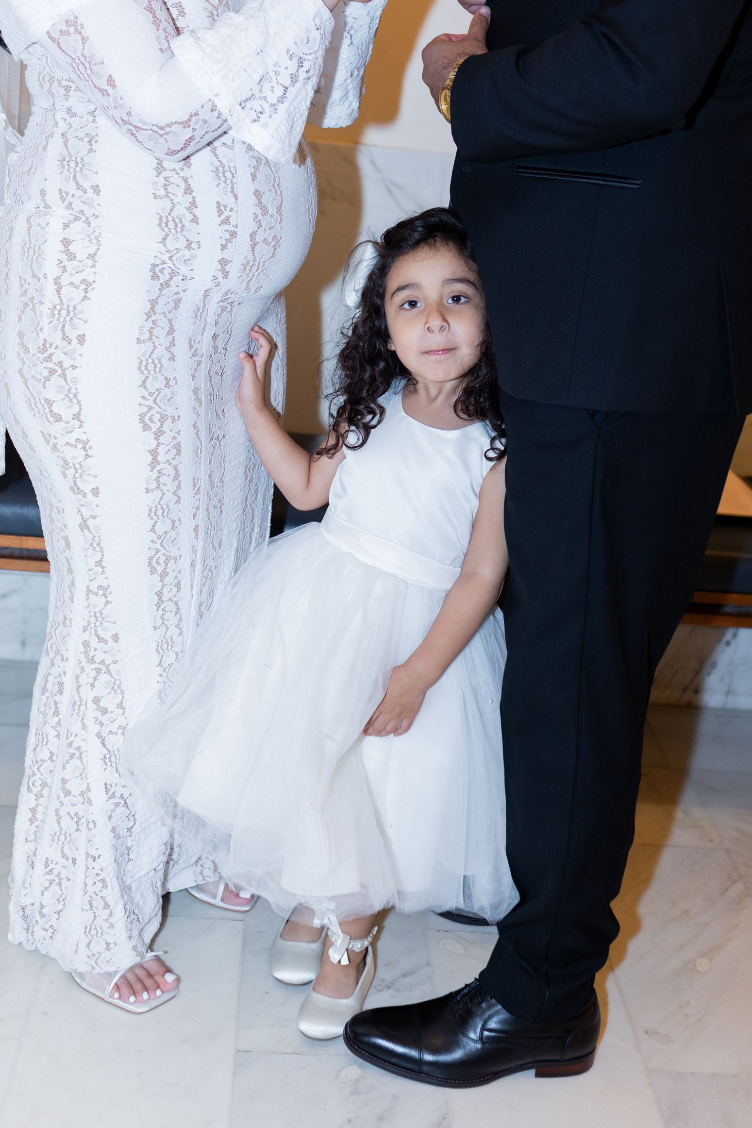 Young flower girl in white tulle dress stands beside bride in lace gown and groom in black tuxedo at wedding.