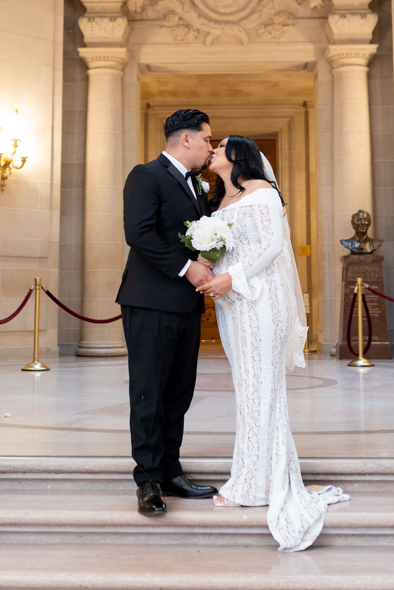 Bride and groom share a kiss in a white lace gown and black tuxedo at San Francisco City Hall.