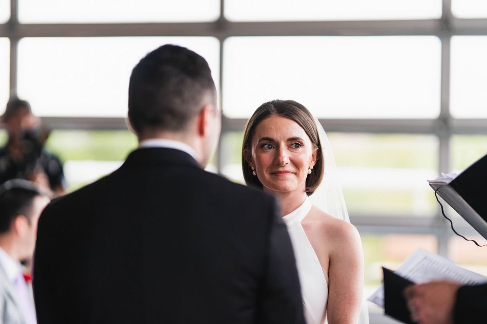 Bride in white halter dress smiling at groom in black suit during wedding ceremony indoors.