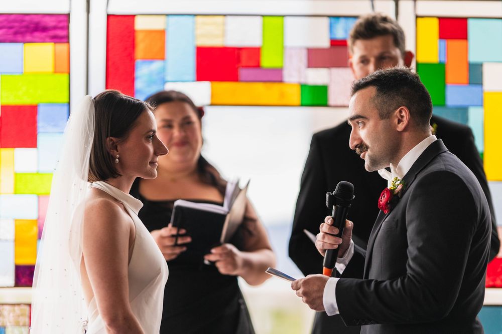 Bride and groom exchange vows at colorful stained glass wedding ceremony with officiant holding book.
