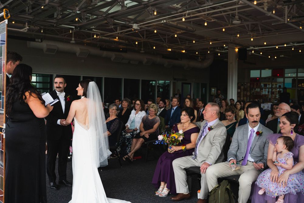 Bride and groom exchange vows at elegant indoor wedding ceremony with string lights and seated guests watching.