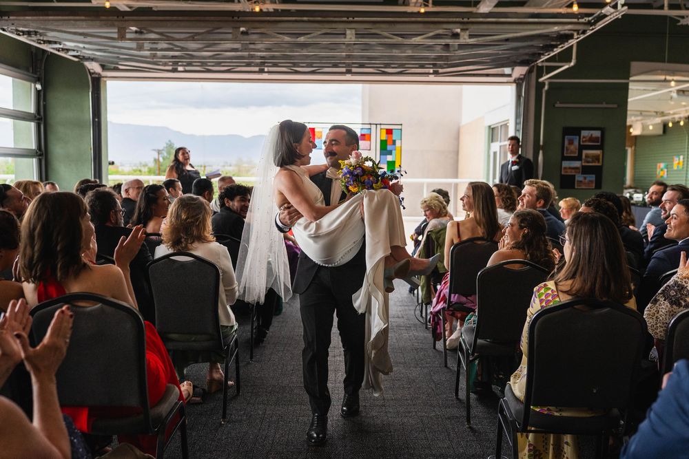 Couple shares first kiss at indoor wedding ceremony with seated guests and mountain views in background.