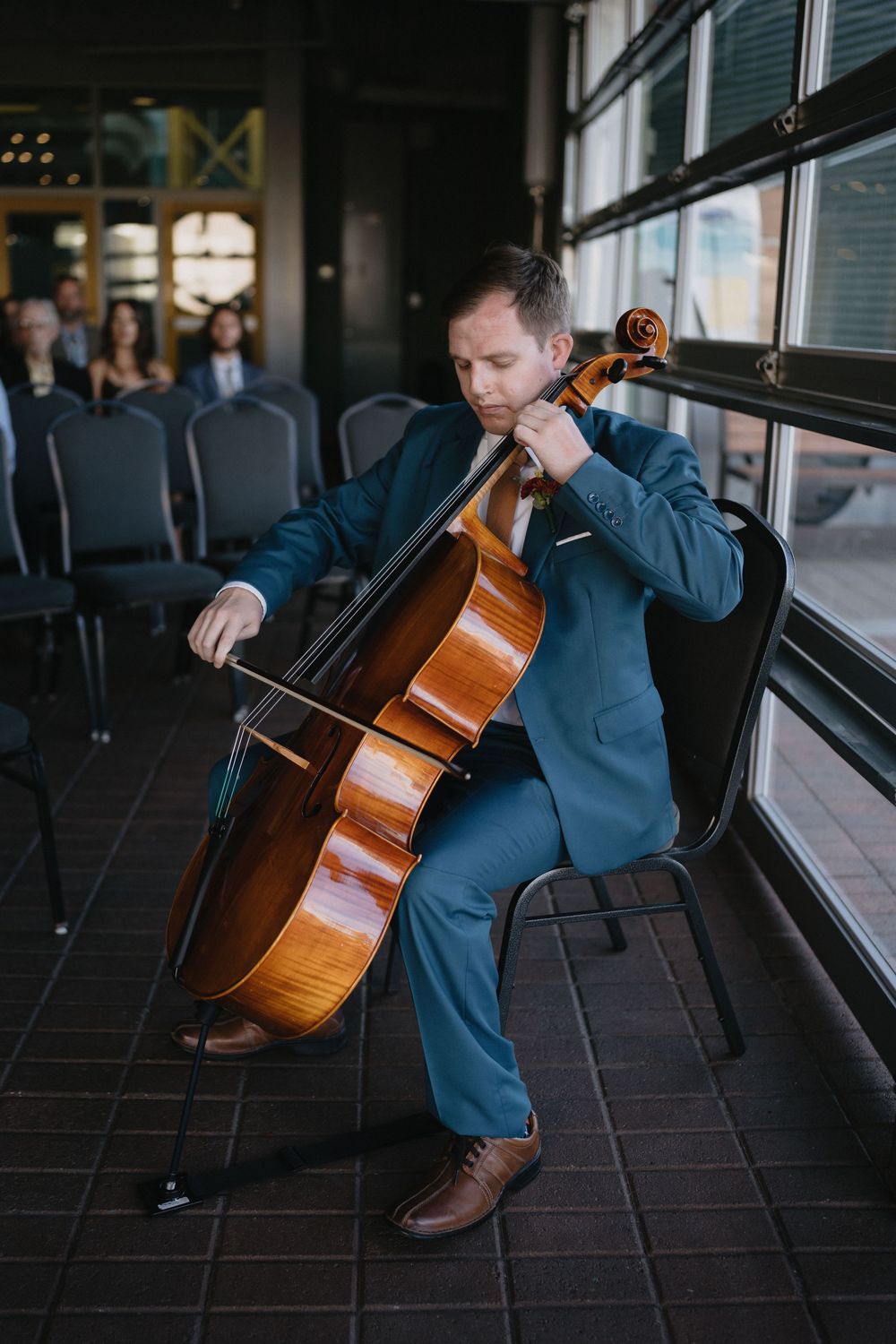 Cellist in blue suit playing cello seated by large windows at an indoor event venue.