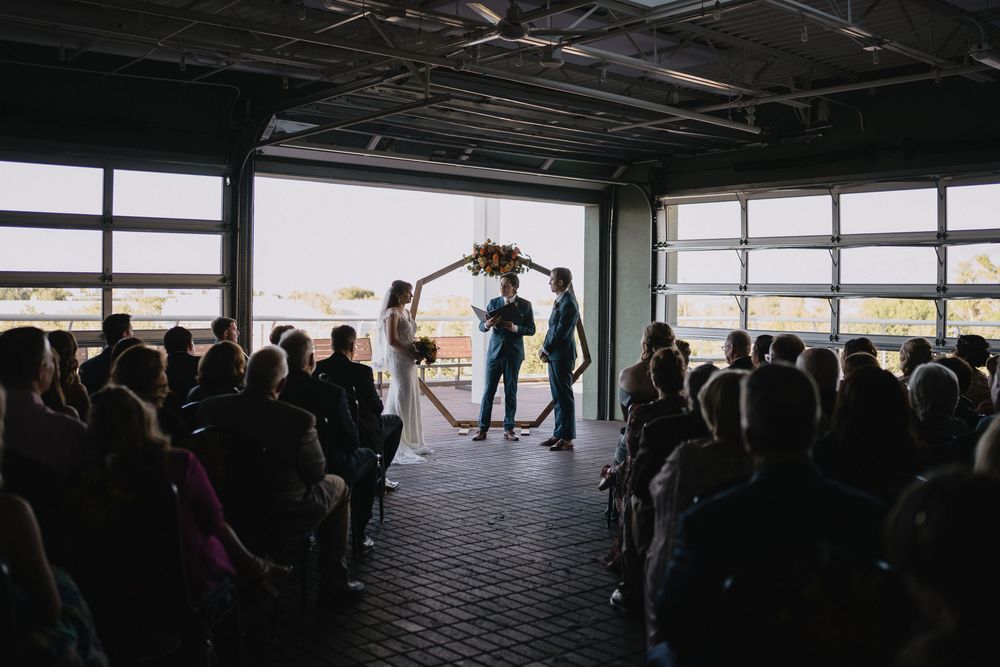 Wedding ceremony in industrial venue with garage doors open, couple holding hands before officiant with seated guests.