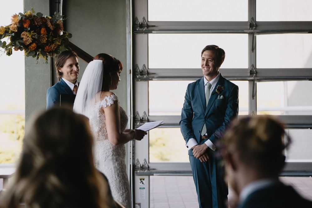 Groom smiling at bride during intimate indoor wedding ceremony with floral arch and large window backdrop.