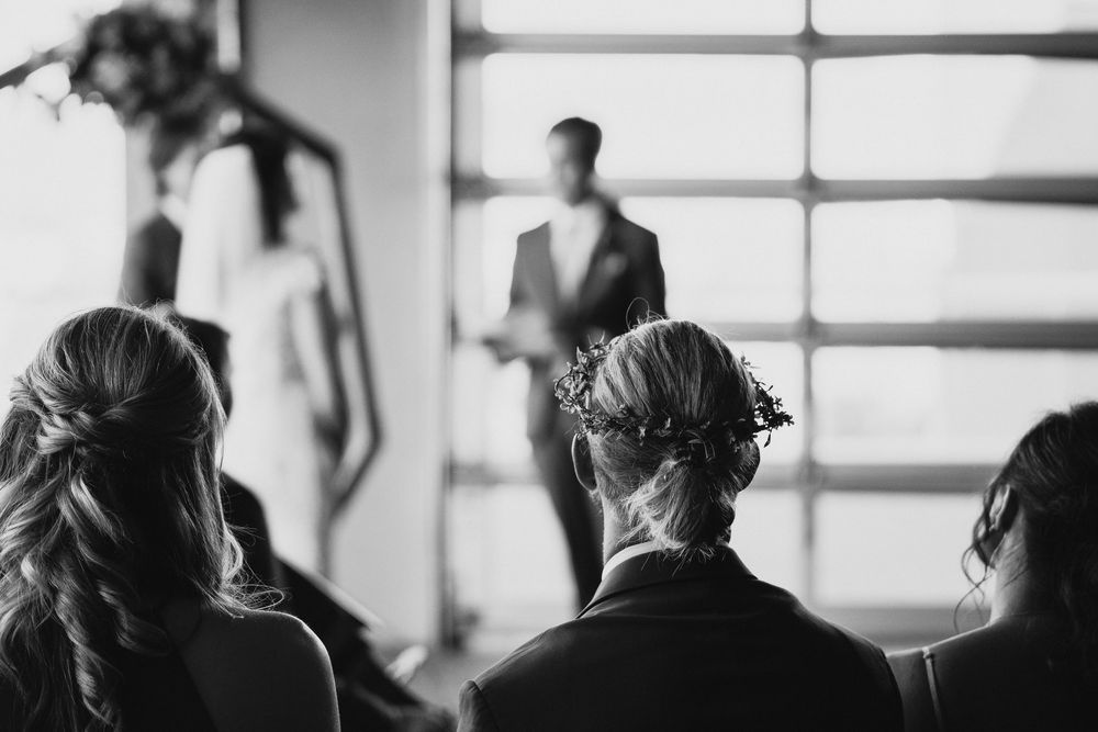 Black and white wedding ceremony photo showing guests watching couple exchange vows near large windows.