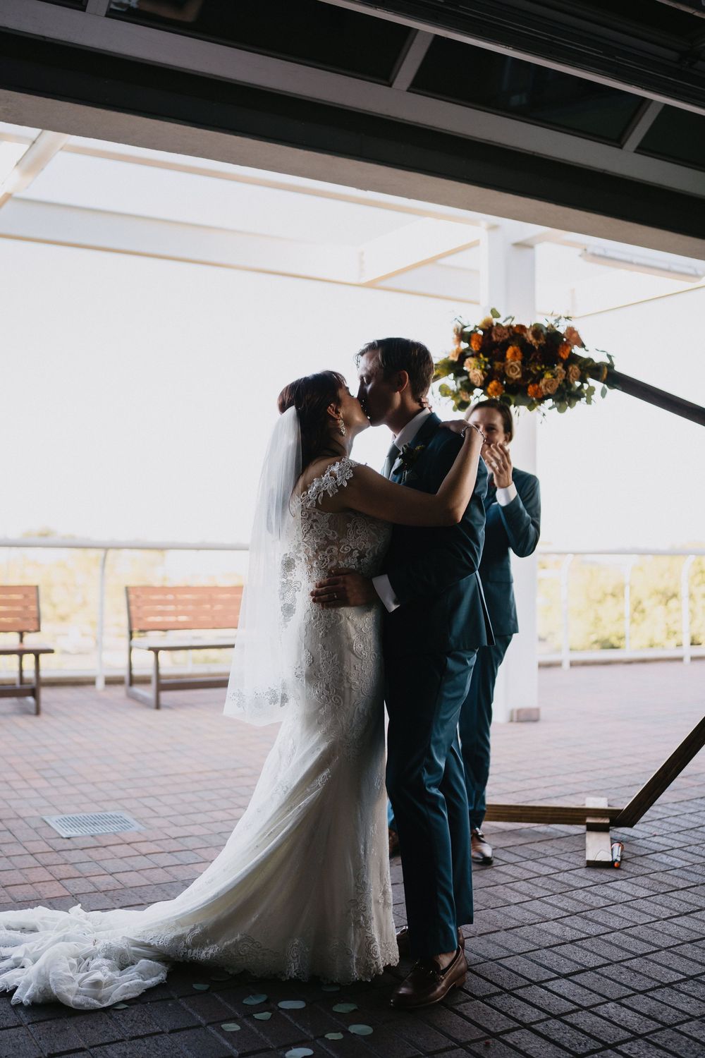 Bride and groom share first kiss at outdoor wedding ceremony, bride in lace gown with veil holding autumn bouquet.