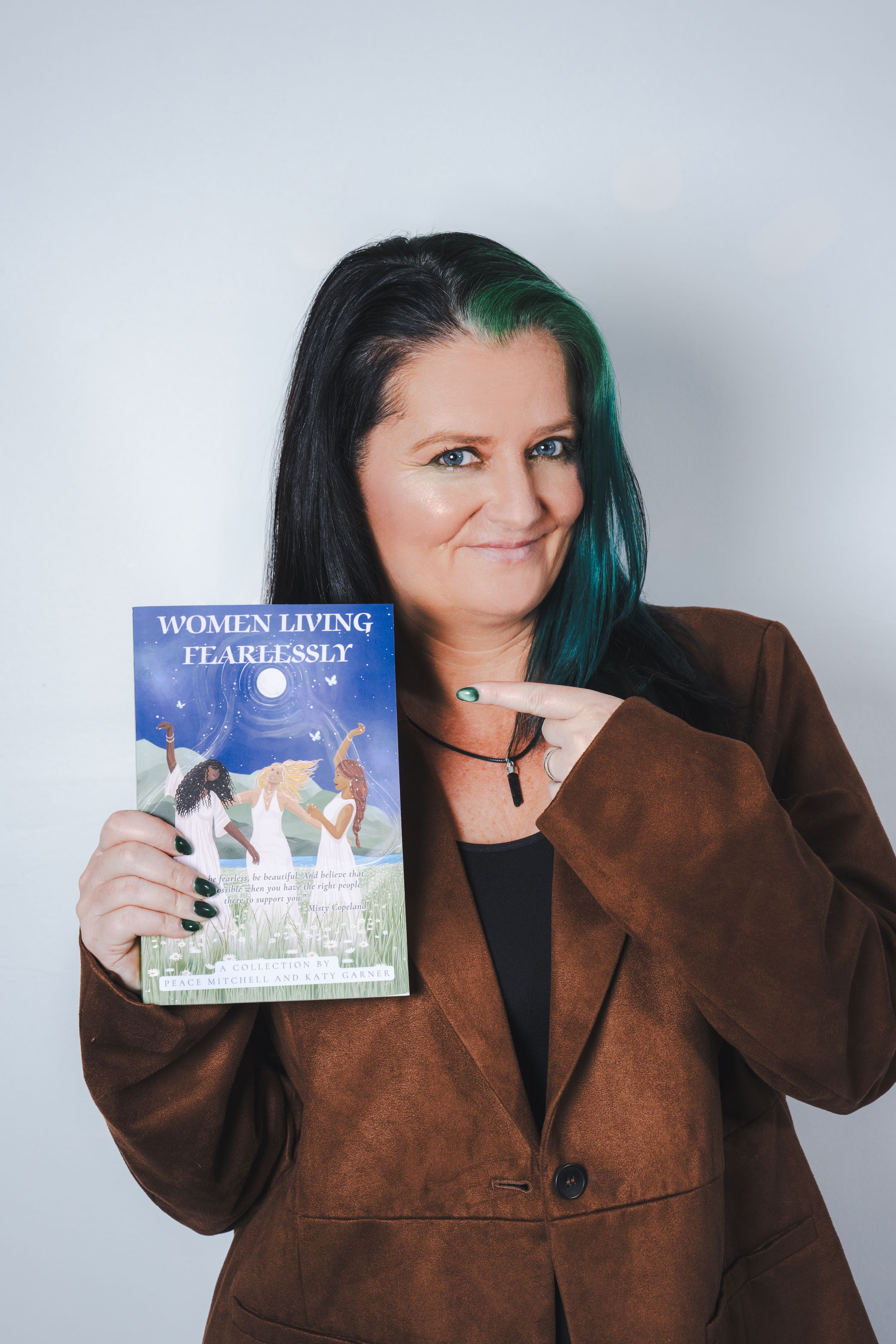 Woman with teal-highlighted hair holding and pointing to book titled Women Living Fearlessly in brown blazer.
