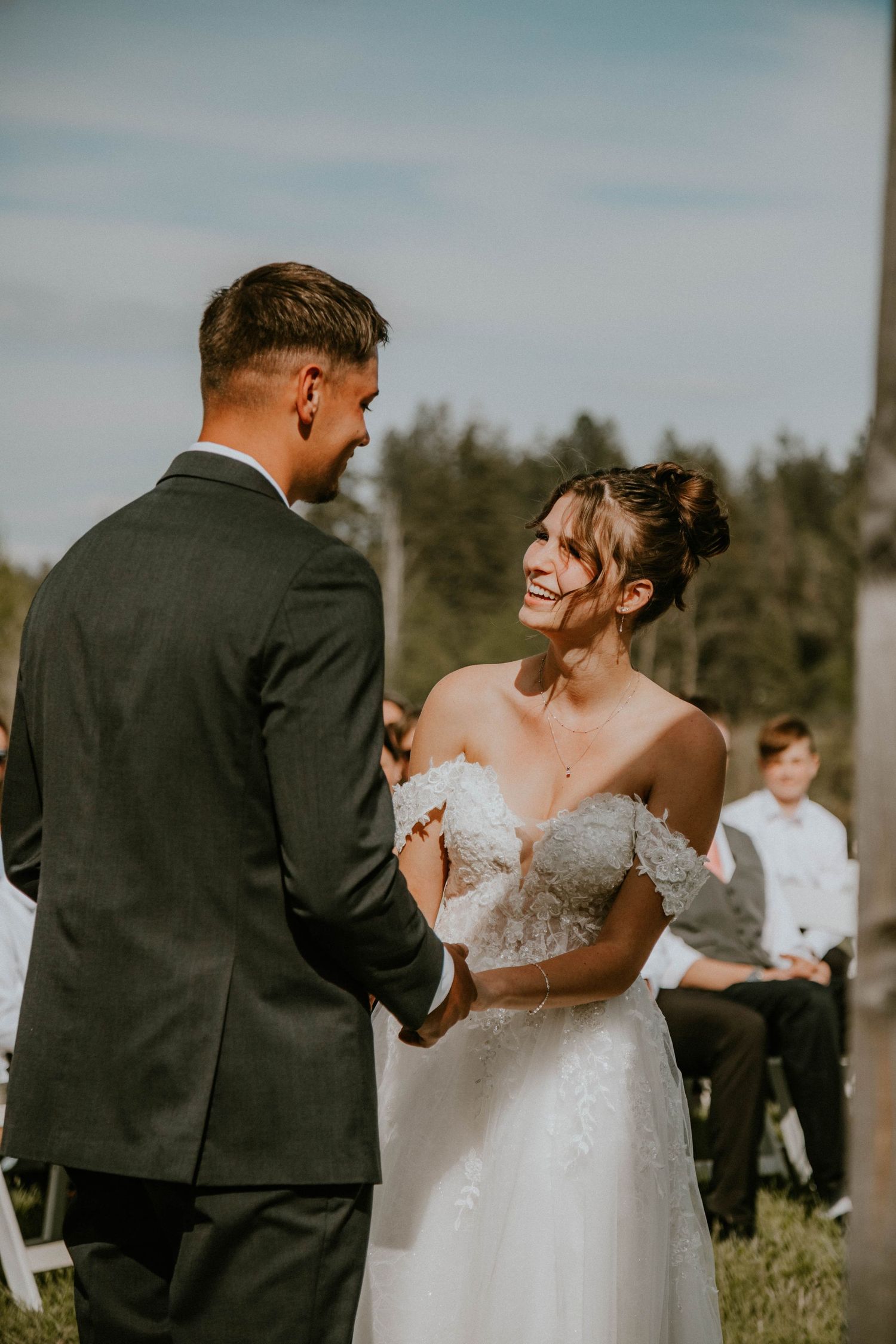 Bride walks down outdoor aisle with escort, holding white bouquet, guests seated on white chairs at sunny backyard wedding.