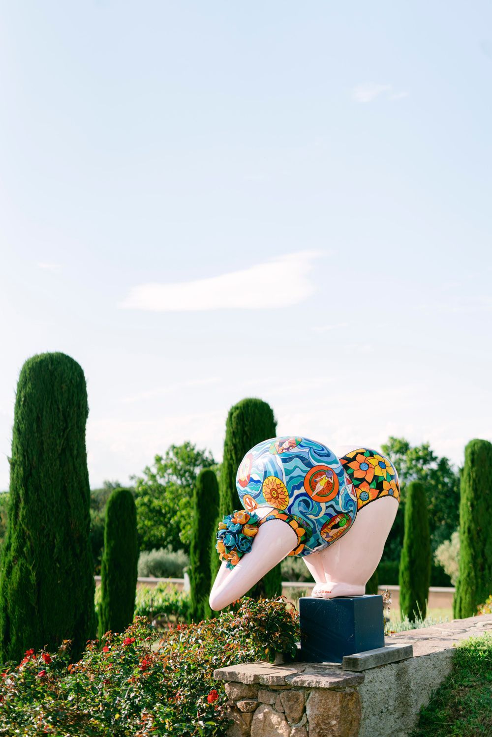 Colorful floral sculpture of a woman's bust in a garden with tall cypress trees under a clear blue sky.