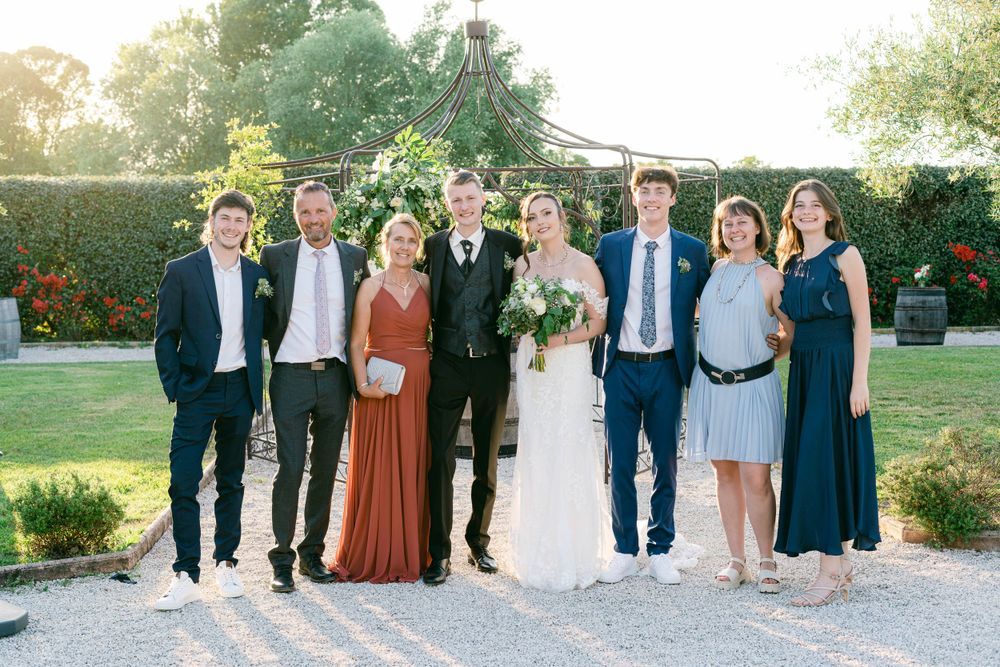 Wedding party of seven posing outdoors in formal attire, bride in white gown holding bouquet, surrounded by guests in navy and rust.