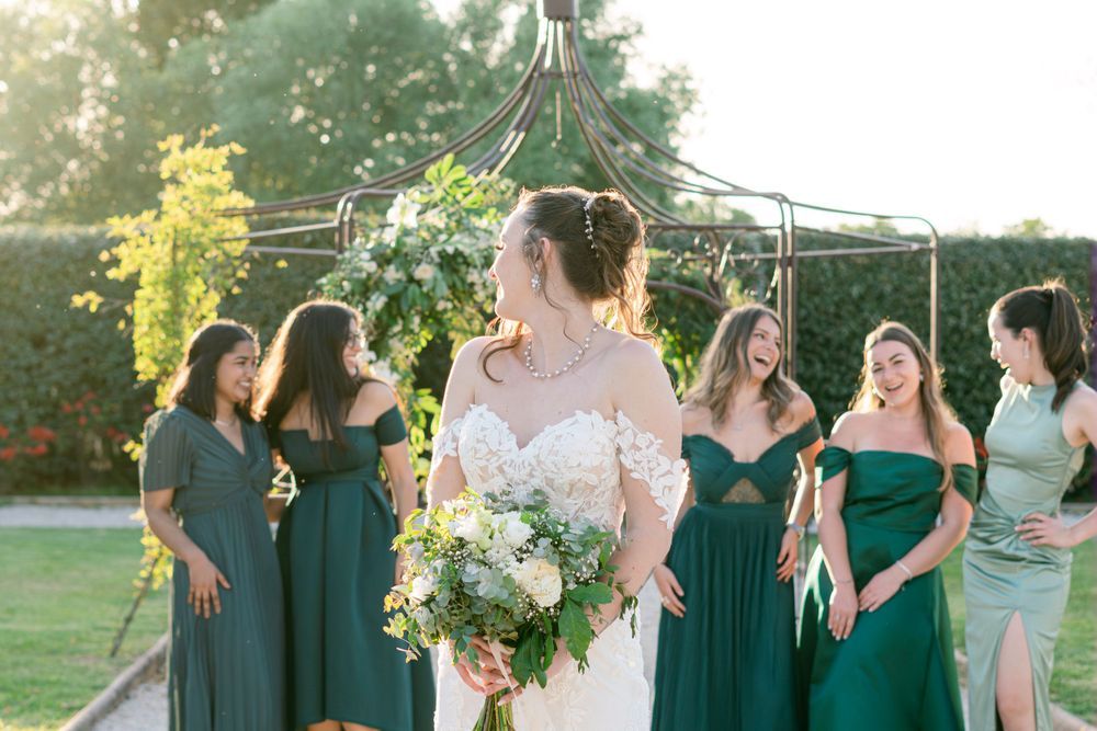 Bride in white gown holds bouquet surrounded by bridesmaids in green dresses outdoors at a sunny wedding ceremony.
