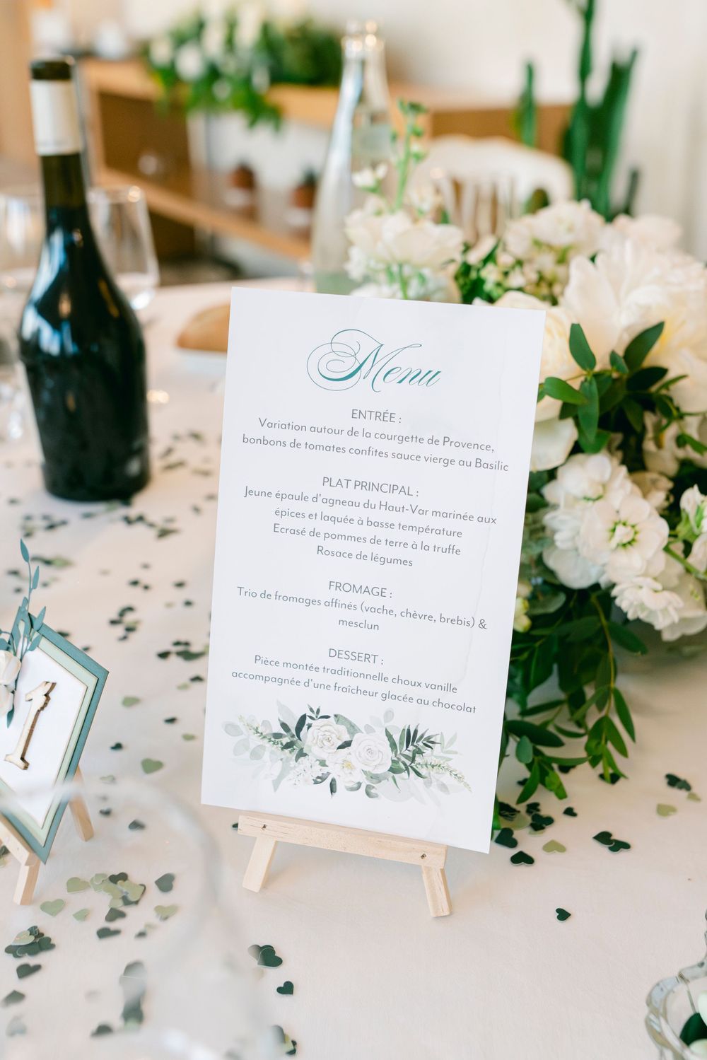 Elegant wedding reception menu card on easel with white floral arrangement and wine bottle on decorated table.