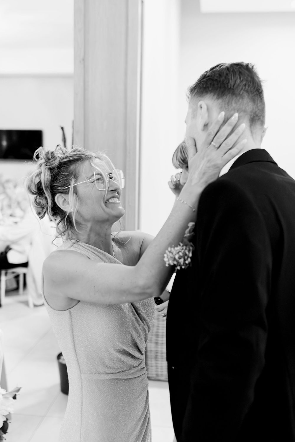 Black and white photo of a smiling woman in glasses tenderly holding a mans face at a wedding reception.
