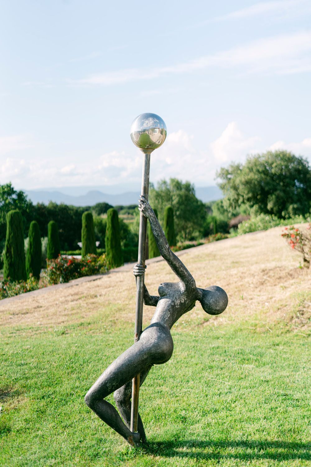 Abstract metal sculpture of a figure holding a reflective sphere on a pole, set in a lush green garden landscape.