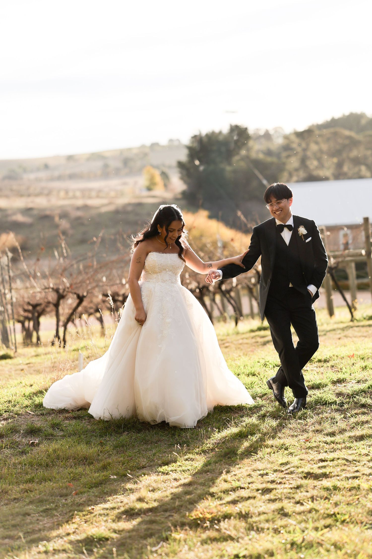 Bride and groom walk hand-in-hand through a sunlit vineyard, she in a flowing white gown, he in a classic black tuxedo.
