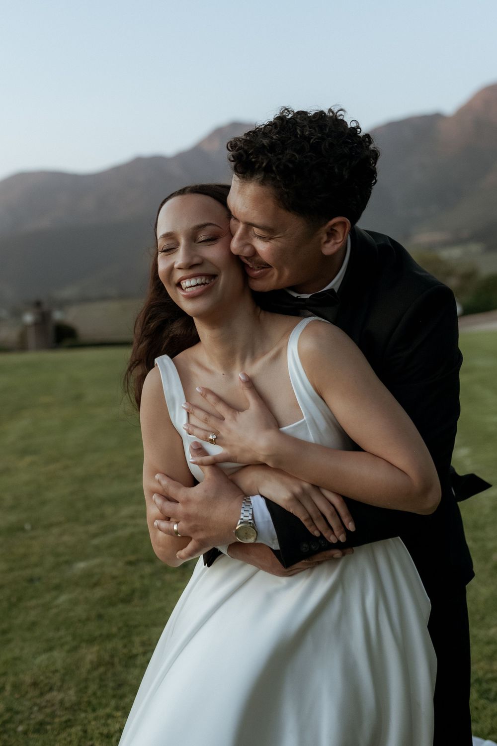 A romantic couple embraces in a grassy field with mountains in the background during a sunset photoshoot.