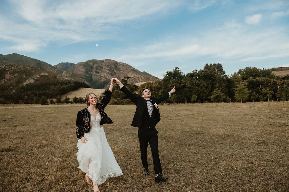 A couple celebrates with raised arms in a scenic mountain field on their wedding day.