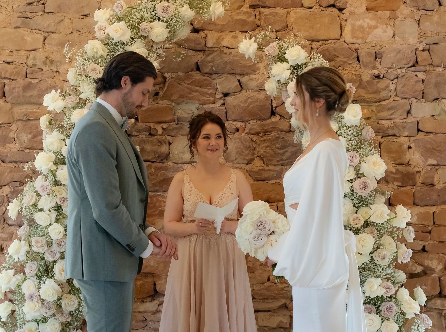 Bride and groom exchange vows beneath a stunning white floral arch officiated by a woman in blush dress.