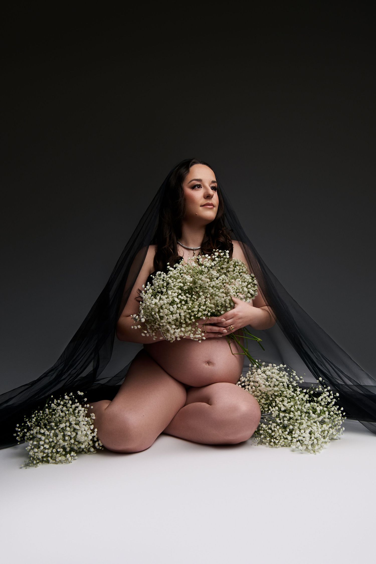 Pregnant woman sitting on floor with white baby's breath flowers, draped in black veil, studio maternity portrait.