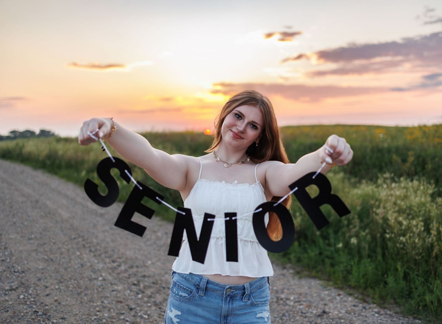 Person holding up black 'SENIOR' letter banner on rural dirt road at sunset with golden sky in background.