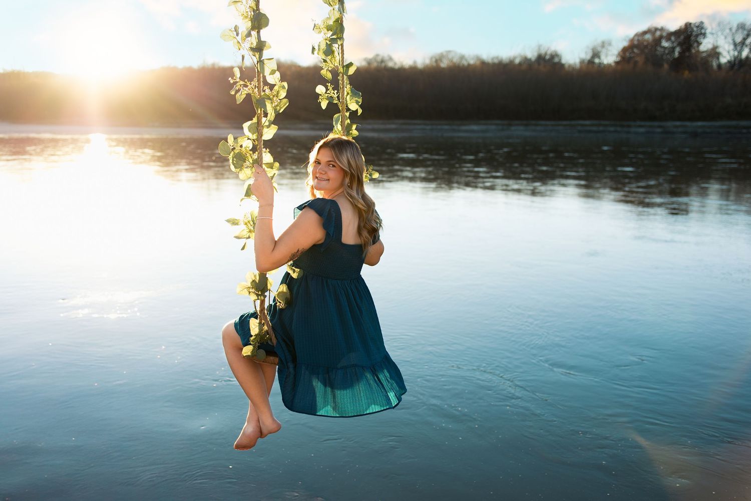 A person swings on a rope swing over a tranquil lake at sunset, creating a silhouette against the golden sky. Senior photography