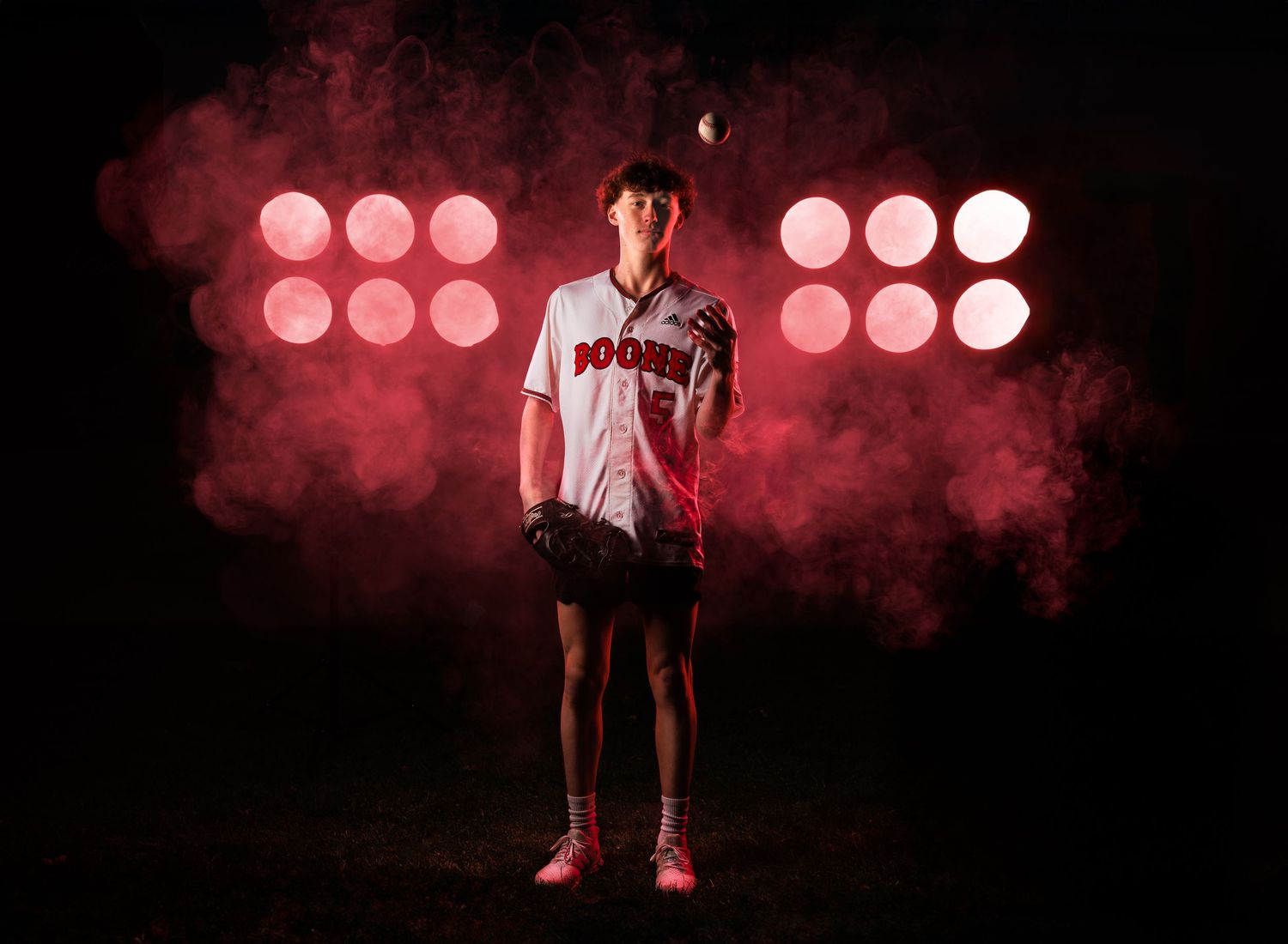 Baseball player stands in dramatic pose against red spotlights and smoke effects in dark studio setting. Senior photography, senior photos