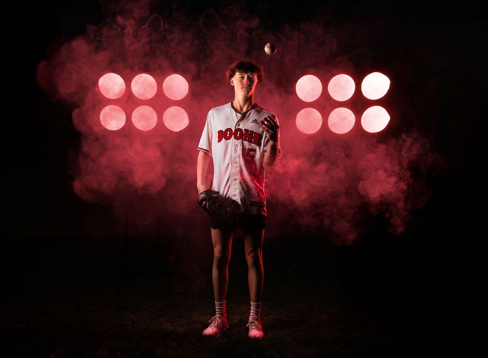 Baseball player stands in dramatic pose against red spotlights and smoke effects in dark studio setting. Senior photography, senior photos