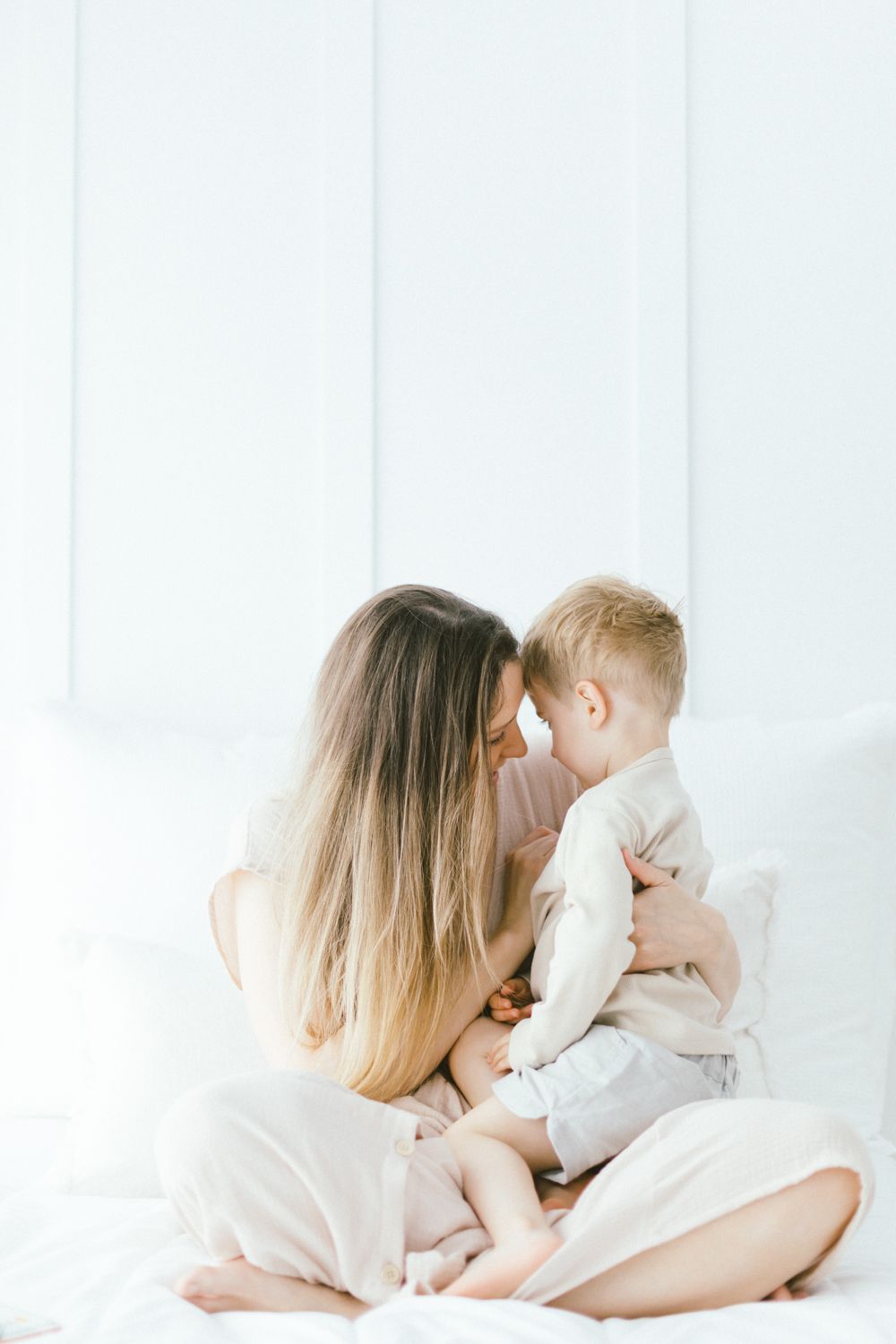 Gentle family cuddles and embraces on white bedding in soft morning light.