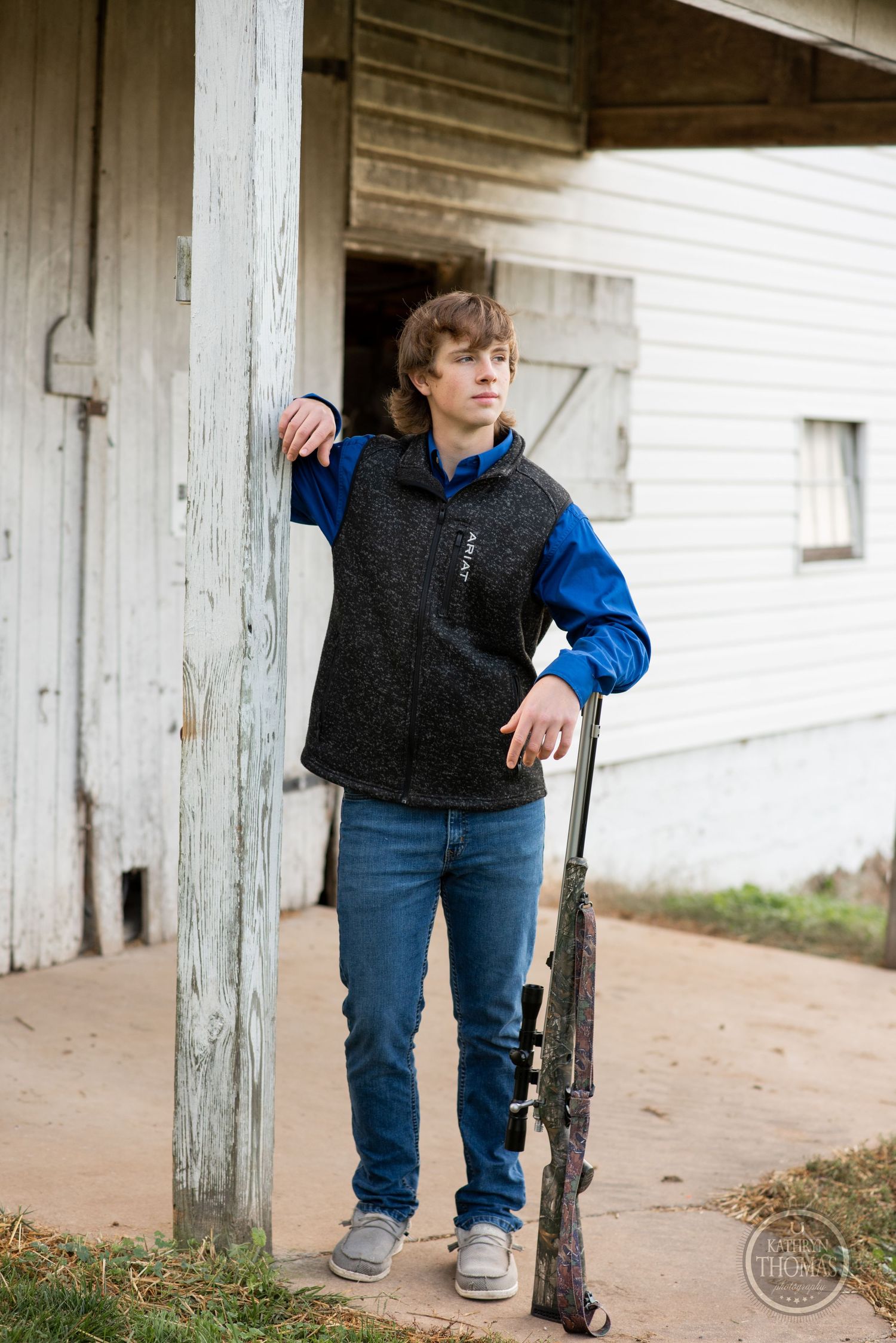 Figure stands by white barn door wearing dark vest and blue sleeves while holding hunting gear.
