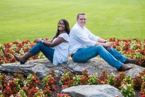 A couple sits back-to-back on a rock wall surrounded by colorful flower beds and lush green lawn.