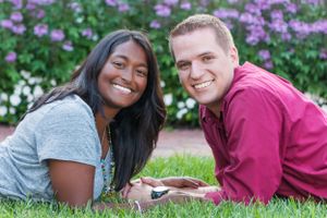 A couple lying in green grass smiling at the camera with purple flowers blooming in the background.