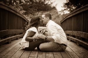 A romantic sepia-toned moment between a couple sitting on a wooden bridge walkway during an engagement photo session.