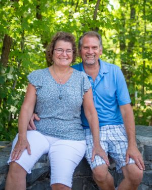 Couple sitting together on stone wall in sunny forest wearing casual summer clothing among green foliage.