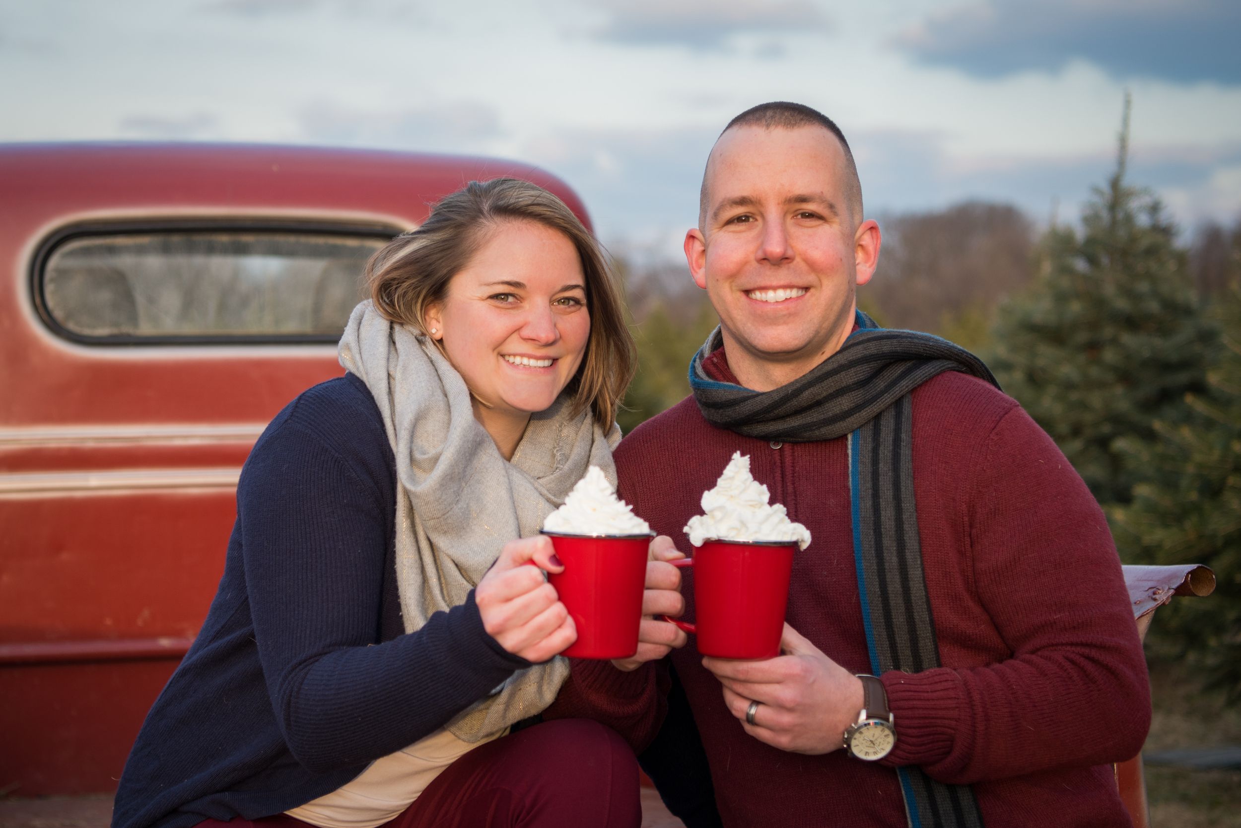 Two people holding red cups with whipped cream outdoors near a vintage red car in evening light.