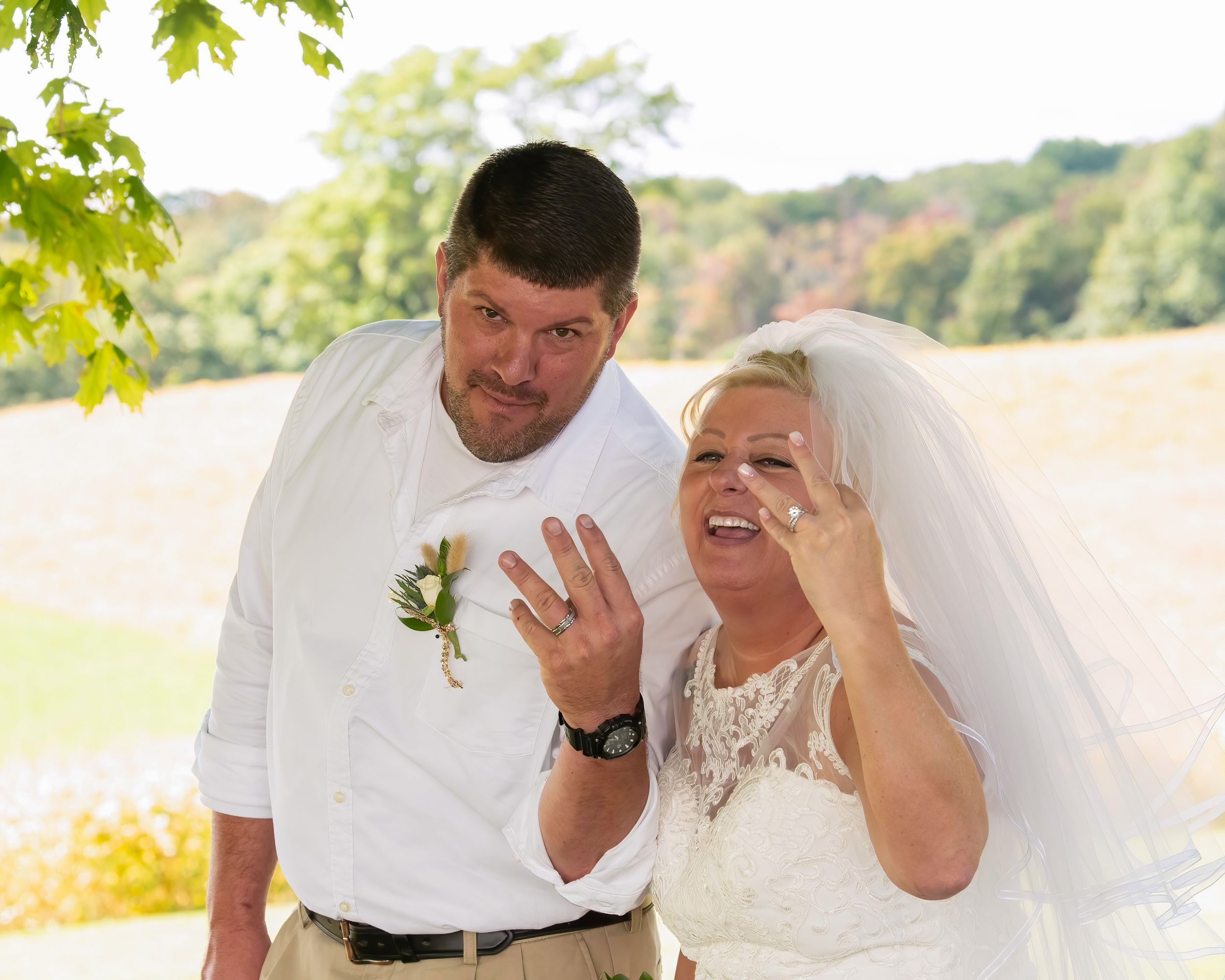 A joyful couple laughs together outdoors during their wedding ceremony while showing off a ring in the sunlight.