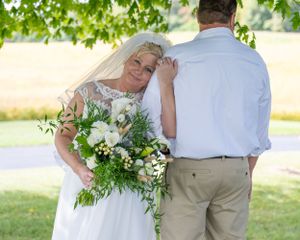 A couple poses outdoors on their wedding day with a lush bouquet of white flowers and greenery.