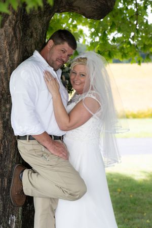 A romantic wedding couple poses together against a tree trunk in an outdoor summer setting.