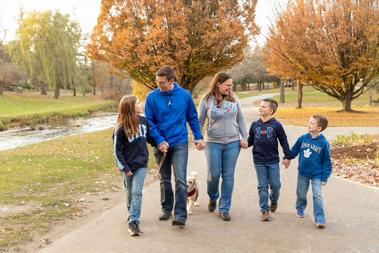 A family photo taken in the fall with all the colourful trees in the background with 3 kids, their parents and a dog.
