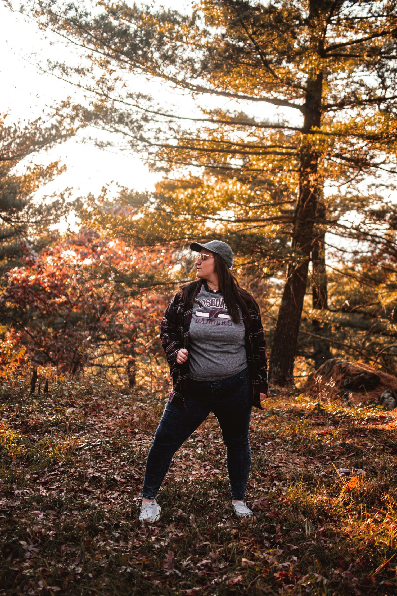 Person in casual wear poses confidently against backdrop of sun-dappled autumn trees.