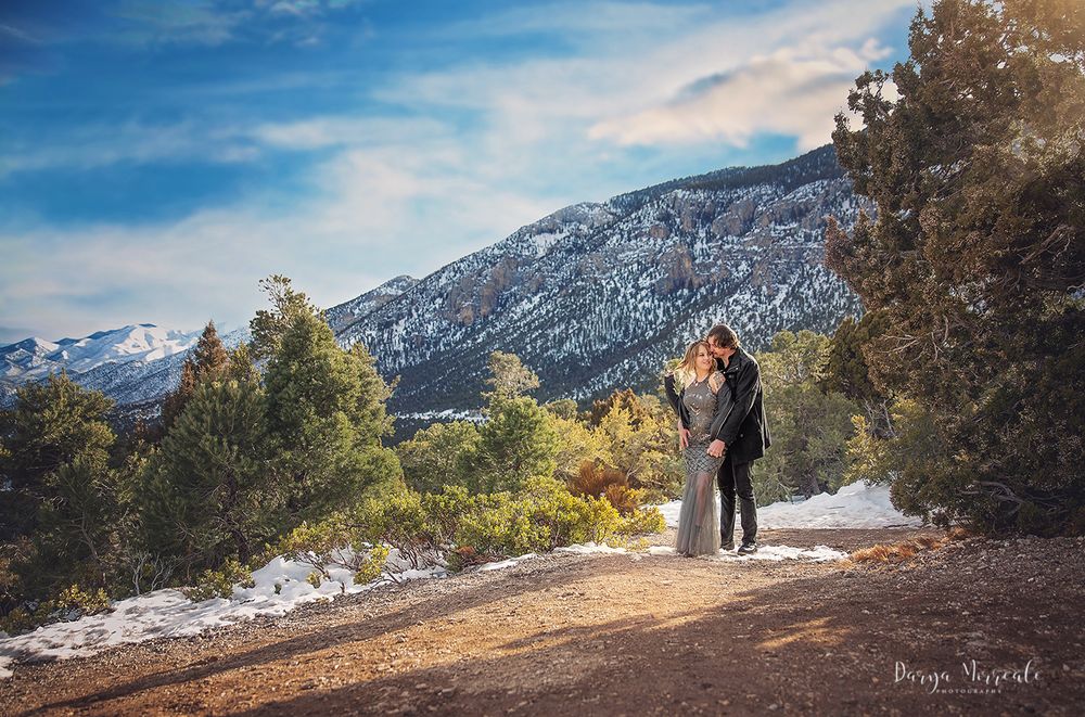 Couple standing on mountain trail surrounded by snow-dusted pines and dramatic peaks.