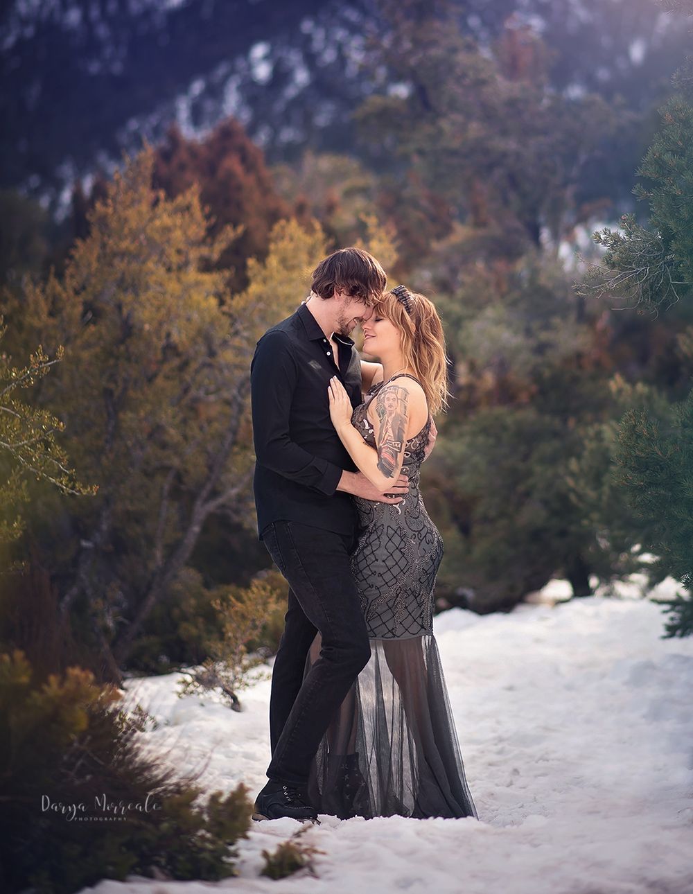 Couple in formal wear embracing on snowy ground with autumn foliage and mountains behind.