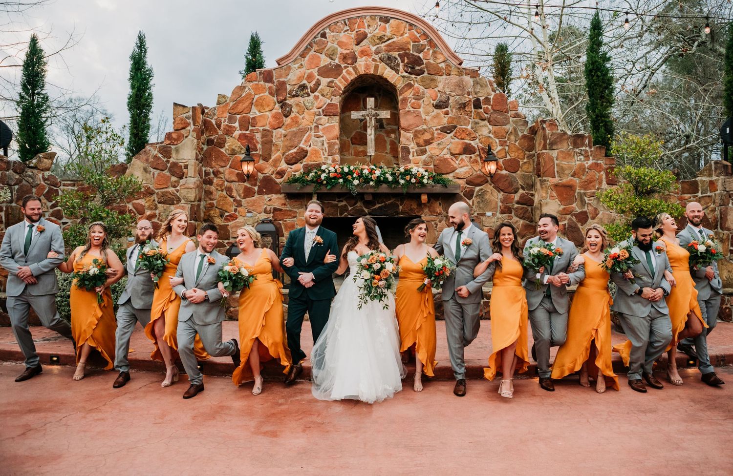 Wedding party in orange dresses and gray suits poses in front of ornate stone archway with cypress trees.