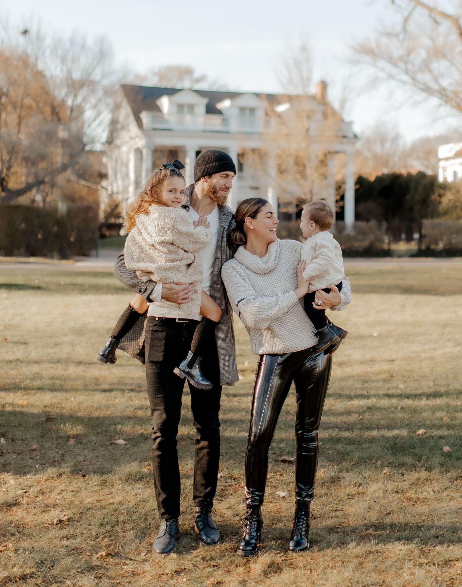 A family of four wearing coordinating cream sweaters poses in front of a historic house.